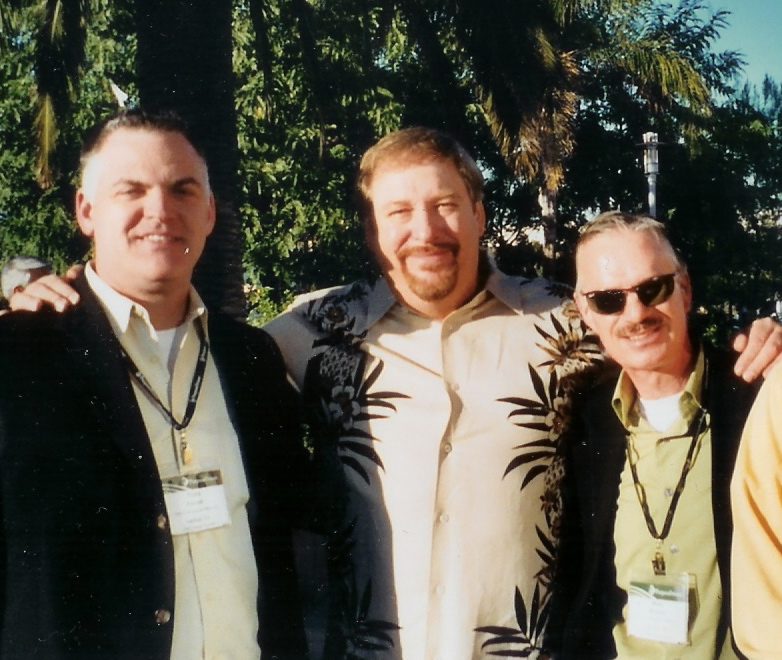Three men smiling outdoors by palm tree