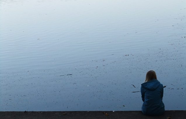 A woman sits on the edge of a lake looking at the water.