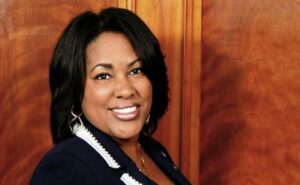A woman in a blue jacket smiling in front of a wooden wall.