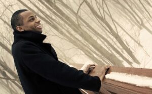 A man in a black coat leaning on a railing in the woods.